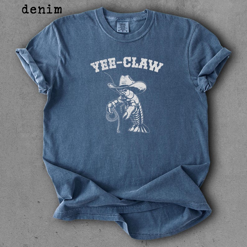 May include: Denim blue t-shirt with rolled sleeves. The shirt features a white graphic of a lobster wearing a cowboy hat and holding a rope, with the text "YEE-CLAW" above the graphic.