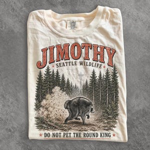 Op de afbeelding: Een cr&egrave;mekleurig t-shirt met een afbeelding van een wasbeer in een bos met de Space Needle op de achtergrond. De tekst luidt JIMOTHY SEATTLE WILDLIFE en DO NOT PET THE ROUND KING. Het shirt heeft een Comfort Colors merklabel in de nek.