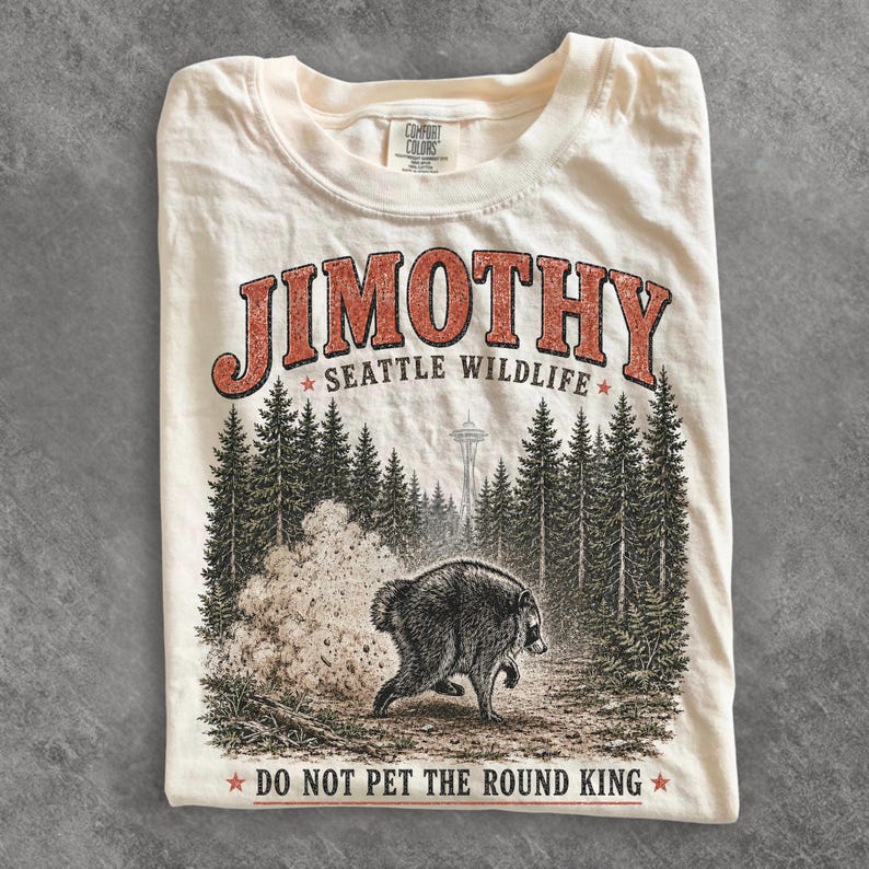 Op de afbeelding: Een cr&egrave;mekleurig t-shirt met een afbeelding van een wasbeer in een bos met de Space Needle op de achtergrond. De tekst luidt JIMOTHY SEATTLE WILDLIFE en DO NOT PET THE ROUND KING. Het shirt heeft een Comfort Colors merklabel in de nek.