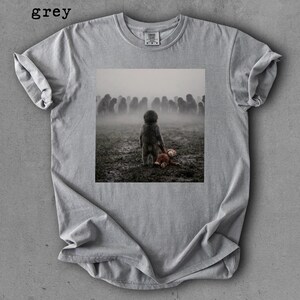 May include: Grey t-shirt with a square graphic of a child holding a stuffed animal in a foggy field with a crowd of figures in the background. The word "grey" is printed at the top left.