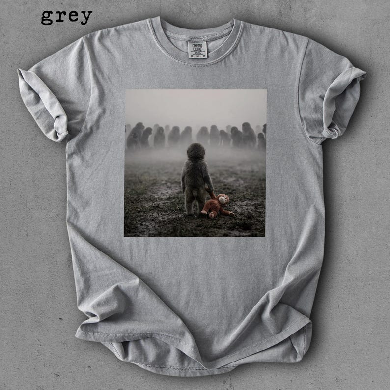 May include: Grey t-shirt with a square graphic of a child holding a stuffed animal in a foggy field with a crowd of figures in the background. The word "grey" is printed at the top left.