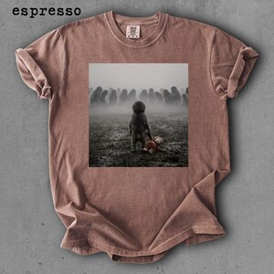 May include: Espresso-colored t-shirt featuring a square image of a child holding a teddy bear, facing a crowd in a foggy field. The shirt has a crew neck and short sleeves. The word "espresso" is printed at the top left.