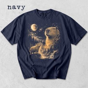 May include: Navy blue t-shirt featuring a detailed illustration of capybaras under a starry night sky with a full moon. The word "navy" is printed at the top left. The design includes a waterfall and forest.