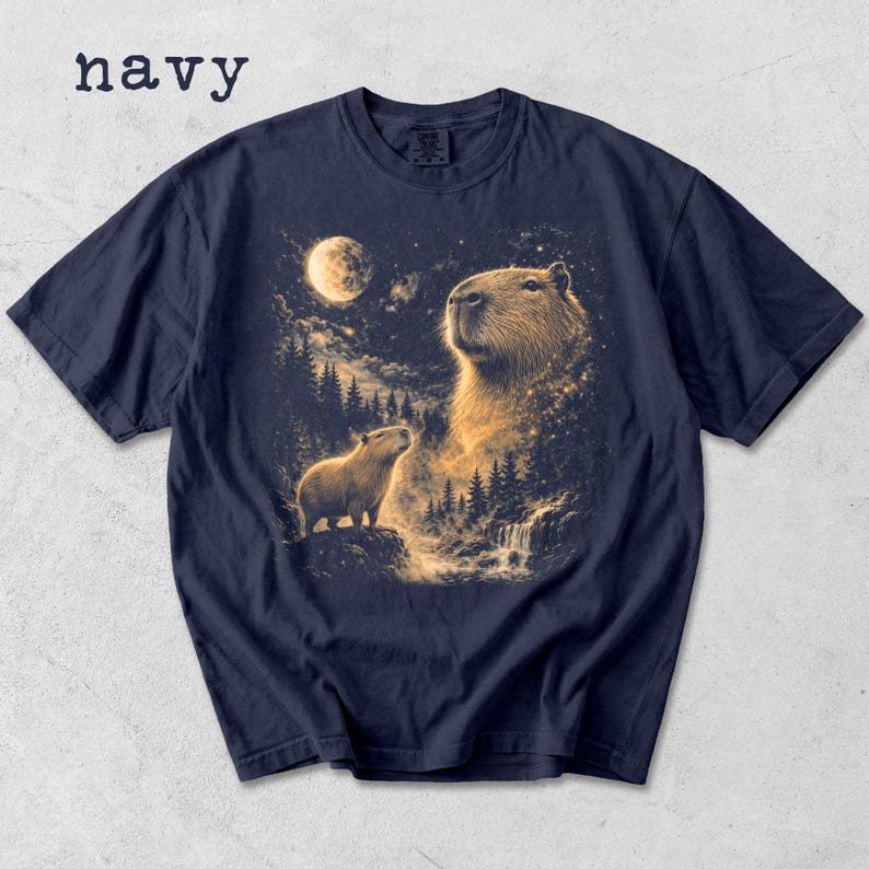 May include: Navy blue t-shirt featuring a detailed illustration of capybaras under a starry night sky with a full moon. The word "navy" is printed at the top left. The design includes a waterfall and forest.