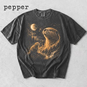 May include: Dark gray t-shirt featuring a detailed illustration of two otters gazing at a moonlit sky. The design includes a forest, waterfall, and the word "pepper" at the top. The artwork is in shades of gold and black.
