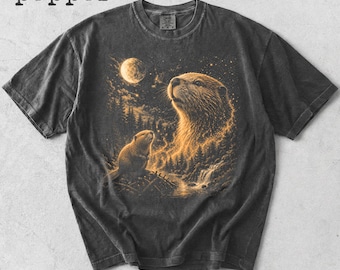 May include: Dark gray t-shirt featuring a detailed illustration of two otters gazing at a moonlit sky. The design includes a forest, waterfall, and the word "pepper" at the top. The artwork is in shades of gold and black.