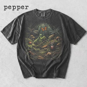 以下が含まれることがあります： 暗い灰色のTシャツ。 昆虫、カマキリ、蝶、トンボなど、詳細な森のシーンが描かれています。 左上に「pepper」の文字がプリントされています。 ヴィンテージ感のあるシャツです。