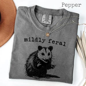 May include: A gray t-shirt with the text "mildly feral" above a black illustration of an opossum. The shirt has a crew neck and short sleeves. A silver necklace with a small pendant is draped across the shirt.