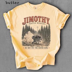 Op de afbeelding: Een botergeel t-shirt met korte mouwen en een afbeelding van een beer in een bos met de Space Needle op de achtergrond. De tekst luidt JIMOTHY SEATTLE WILDLIFE en DO NOT PET THE ROUND KING in rode en zwarte letters.