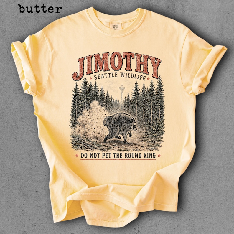 Op de afbeelding: Een botergeel t-shirt met korte mouwen en een afbeelding van een beer in een bos met de Space Needle op de achtergrond. De tekst luidt JIMOTHY SEATTLE WILDLIFE en DO NOT PET THE ROUND KING in rode en zwarte letters.