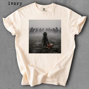 May include: Ivory-colored t-shirt featuring a square image of a child standing in a field with a teddy bear. The child faces away from the viewer, with a crowd of indistinct figures in the background, shrouded in mist.