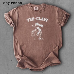 May include: Espresso-colored t-shirt featuring a white graphic of a lobster wearing a cowboy hat and holding a lasso. The text "YEE-CLAW" is above the graphic. The shirt has a crew neck and short sleeves.