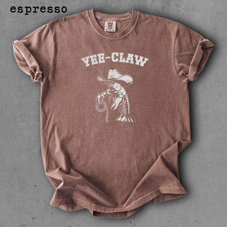 May include: Espresso-colored t-shirt featuring a white graphic of a lobster wearing a cowboy hat and holding a lasso. The text "YEE-CLAW" is above the graphic. The shirt has a crew neck and short sleeves.