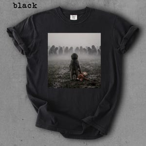 May include: Black t-shirt with a printed image of a child standing in a muddy field, holding a teddy bear. The background features a crowd of indistinct figures in a foggy environment. The word "black" is printed at the top left.