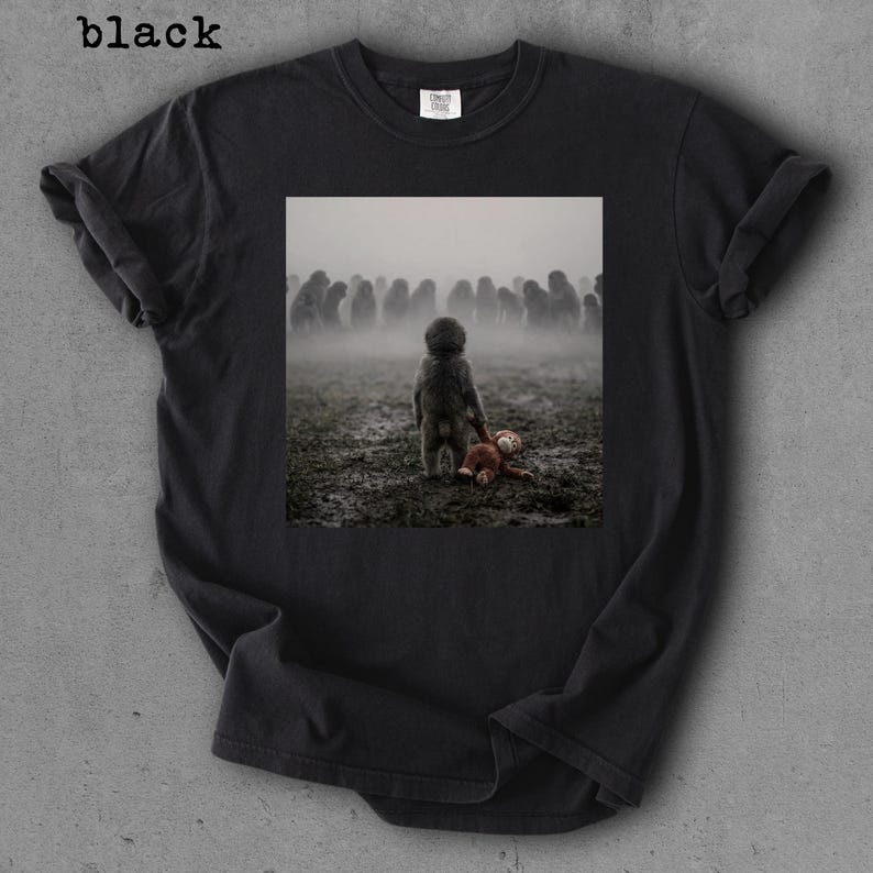 May include: Black t-shirt with a printed image of a child standing in a muddy field, holding a teddy bear. The background features a crowd of indistinct figures in a foggy environment. The word "black" is printed at the top left.