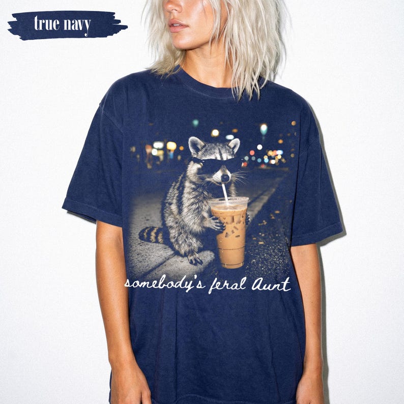 May include: A true navy short-sleeve t-shirt featuring a graphic of a raccoon wearing sunglasses and holding an iced coffee. The text "somebody's feral Aunt" is printed in white script below the raccoon, with "true navy" in the top corner.