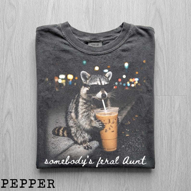 May include: A charcoal gray t-shirt featuring a graphic of a raccoon wearing sunglasses and holding an iced coffee. The text "somebody's feral Aunt" is printed in white script at the bottom. The shirt has a crew neckline and a vintage-style wash.