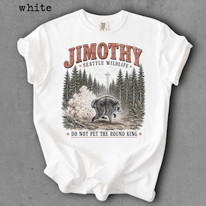 Op de afbeelding: Een wit t-shirt met korte mouwen en een grafische print van een ronde beer in een bos met de Space Needle op de achtergrond. De tekst luidt JIMOTHY SEATTLE WILDLIFE en DO NOT PET THE ROUND KING in rode en zwarte letters.