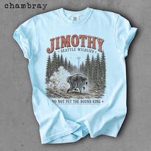 Op de afbeelding: Een lichtblauw t-shirt met een grafische print van een beer in een bos met de Space Needle op de achtergrond. De tekst luidt JIMOTHY SEATTLE WILDLIFE en DO NOT PET THE ROUND KING. Het woord chambray staat in de linkerbovenhoek.