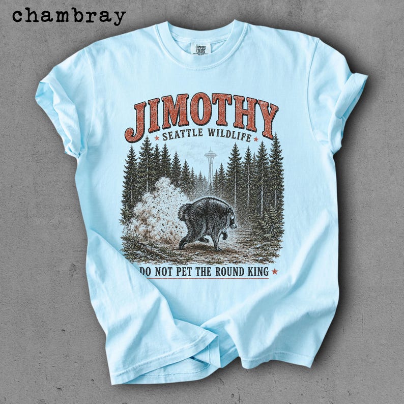 Op de afbeelding: Een lichtblauw t-shirt met een grafische print van een beer in een bos met de Space Needle op de achtergrond. De tekst luidt JIMOTHY SEATTLE WILDLIFE en DO NOT PET THE ROUND KING. Het woord chambray staat in de linkerbovenhoek.