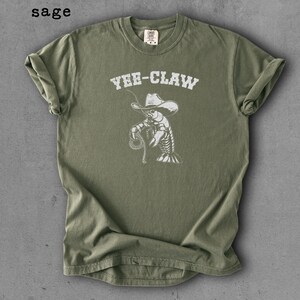 May include: Sage green t-shirt with the text "Yee-Claw" above a white illustration of a cowboy lobster holding a lasso. The shirt has short sleeves and a crew neck.