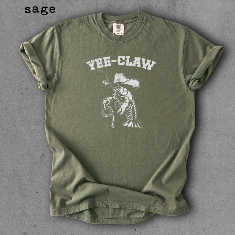May include: Sage green t-shirt with the text "Yee-Claw" above a white illustration of a cowboy lobster holding a lasso. The shirt has short sleeves and a crew neck.
