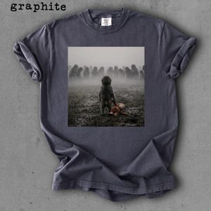 May include: Graphite-colored t-shirt featuring a square image of a child holding a stuffed animal in a foggy field with a crowd of people in the background. The word "graphite" is printed at the top left.