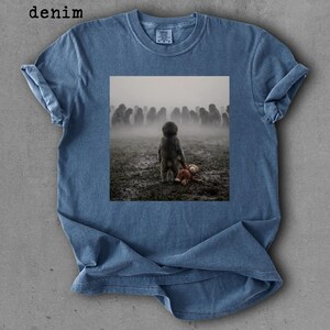 May include: Denim blue t-shirt with a square graphic. The graphic depicts a child holding a teddy bear, facing a crowd in a foggy field. The word "denim" is printed in the upper left corner.