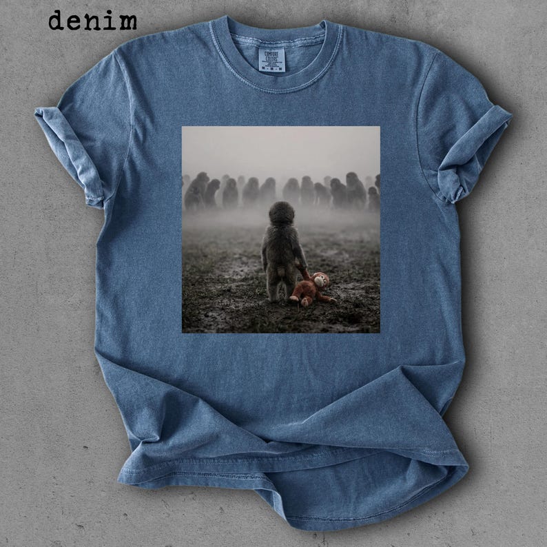 May include: Denim blue t-shirt with a square graphic. The graphic depicts a child holding a teddy bear, facing a crowd in a foggy field. The word "denim" is printed in the upper left corner.