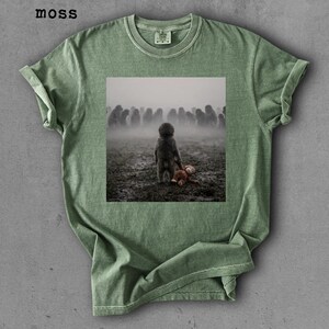 May include: Moss green t-shirt featuring a square graphic of a child with a stuffed animal, facing a crowd in a foggy field. The shirt has a crew neck and rolled-up sleeves. The word "moss" is printed in the upper left corner.