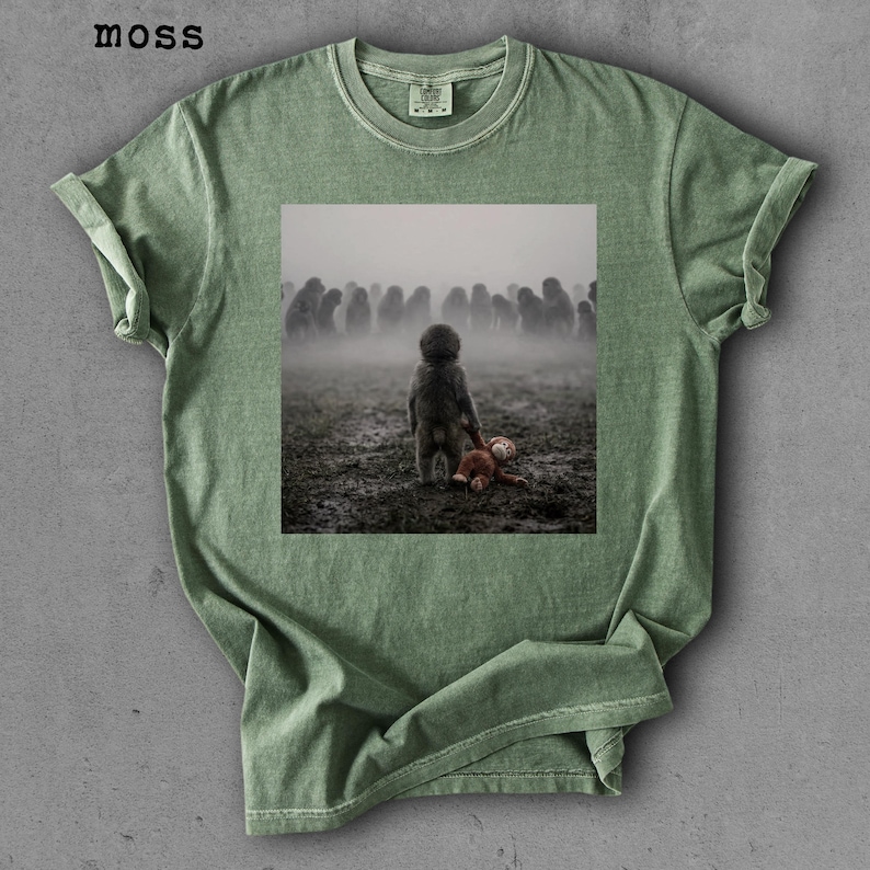 May include: Moss green t-shirt featuring a square graphic of a child with a stuffed animal, facing a crowd in a foggy field. The shirt has a crew neck and rolled-up sleeves. The word "moss" is printed in the upper left corner.