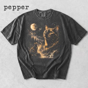 May include: A charcoal grey short-sleeve t-shirt featuring a graphic print of a cat looking up at a full moon in a starry night sky above a forest and waterfall. The word pepper is printed in black text in the upper left corner of the image.