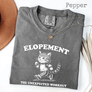 May include: Pepper-colored t-shirt with the word "ELOPEMENT" above a cartoon cat in athletic wear. The text "THE UNEXPECTED WORKOUT" is below the cat. A silver necklace with a pendant is on the shirt.