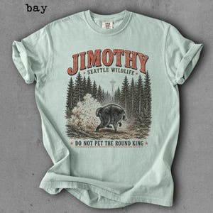 Op de afbeelding: Een lichtgroen T-shirt met korte mouwen en een afbeelding van een beer in een bos met de Space Needle op de achtergrond. De tekst JIMOTHY SEATTLE WILDLIFE en DO NOT PET THE ROUND KING staat in rode en zwarte letters op de voorzijde.