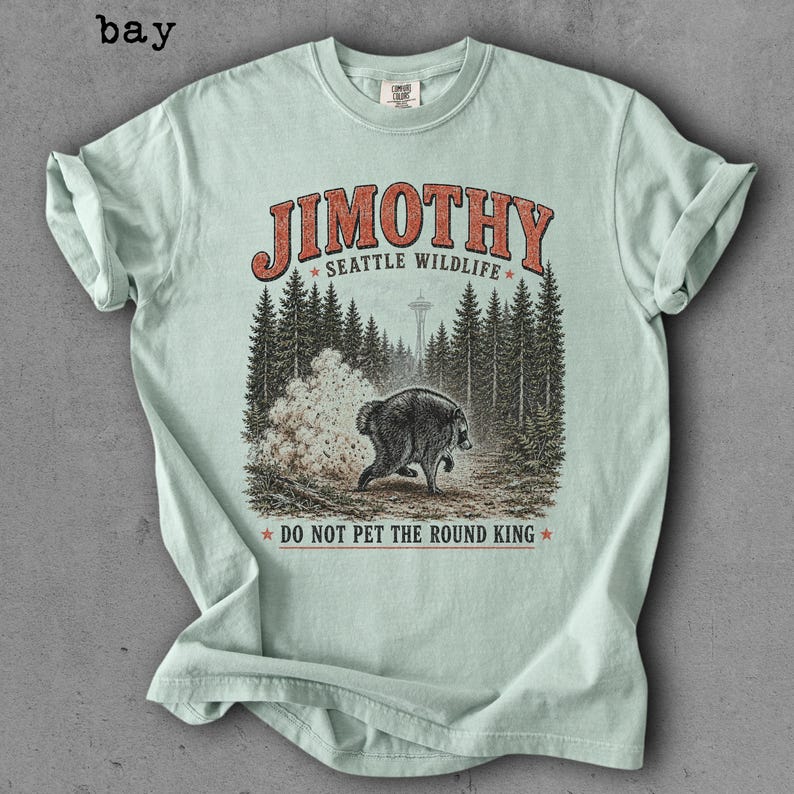 Op de afbeelding: Een lichtgroen T-shirt met korte mouwen en een afbeelding van een beer in een bos met de Space Needle op de achtergrond. De tekst JIMOTHY SEATTLE WILDLIFE en DO NOT PET THE ROUND KING staat in rode en zwarte letters op de voorzijde.