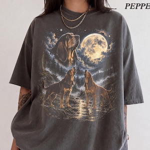 Może przedstawiać: Ciemnoszary t-shirt z nadrukiem graficznym przedstawiającym trzy psy gończe w lesie pod dużą pełnią księżyca. W prawym górnym rogu znajduje się napis "PEPPER". Koszulka ma krótkie rękawy i okrągły dekolt.