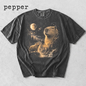 Peut inclure: T-shirt gris foncé avec une illustration de capybara sous la pleine lune. Le motif comprend un grand capybara avec un plus petit, une cascade et des arbres. Le mot "pepper" est imprimé en haut à gauche.