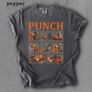 Peut inclure: T-shirt gris fonc&eacute; avec des manches retrouss&eacute;es. Le mot "PUNCH" en lettres orange est imprim&eacute; au-dessus d'une grille de singes de dessins anim&eacute;s. Les singes sont dans diverses poses, certains se c&acirc;linant et d'autres allong&eacute;s.