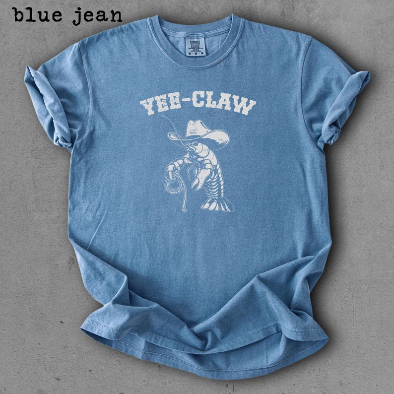 May include: Light blue t-shirt with the text "YEE-CLAW" above a white illustration of a cowboy lobster holding a lasso. The shirt has a crew neck and short sleeves. The words "blue jean" are in the top left corner.