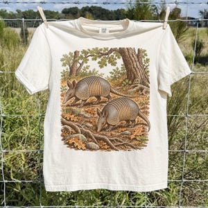 May include: A cream-coloured short-sleeve t-shirt featuring a detailed graphic print of two armadillos in a forest setting with trees, roots, and fallen leaves. The shirt has a crew neckline and a label at the inner collar area.