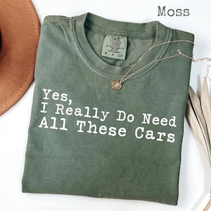 May include: A moss green t-shirt with the text "Yes, I Really Do Need All These Cars" in white. A gold necklace with a pendant rests on the shirt. The shirt is folded and displayed with a brown hat and dried plant.