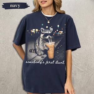 May include: A navy blue short-sleeve t-shirt featuring a graphic of a raccoon wearing black sunglasses and holding a plastic cup with a straw. The text "somebody's feral Aunt" is printed in white cursive script below the raccoon image.