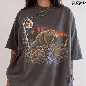 May include: A charcoal grey short-sleeve t-shirt featuring a graphic print of three beavers in a forest setting at night. The scene includes a full moon, a starry sky, and a glowing orange horizon. The word PEPPER is printed in the top right corner.