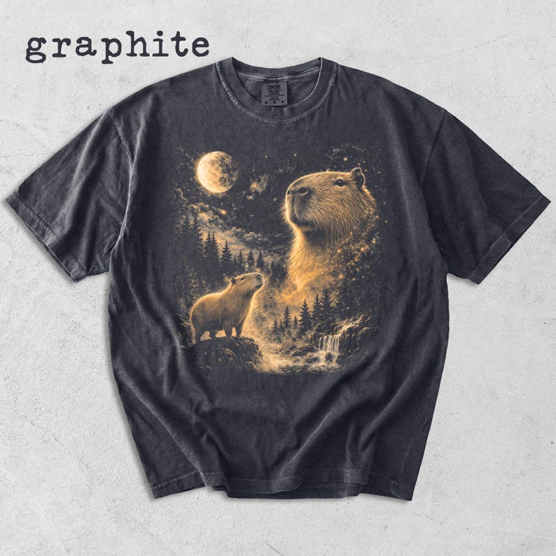 May include: Graphite-colored t-shirt featuring a detailed illustration of two capybaras under a full moon. The design includes a waterfall, trees, and a starry night sky, with the word "graphite" at the top.