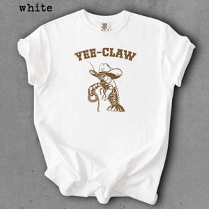 May include: White t-shirt with a brown graphic of a lobster wearing a cowboy hat and holding a rope. The text "YEE-CLAW" is above the lobster. The word "white" is in the top left corner.
