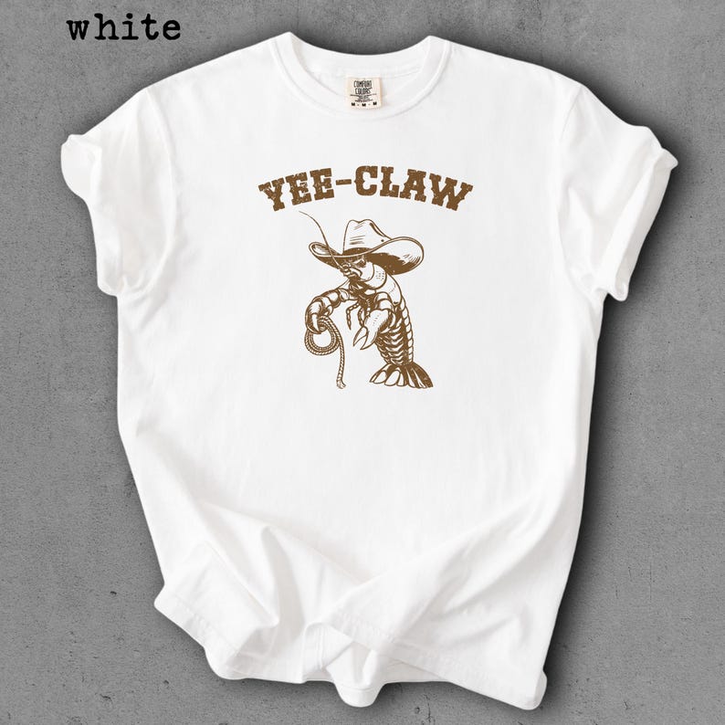 May include: White t-shirt with a brown graphic of a lobster wearing a cowboy hat and holding a rope. The text "YEE-CLAW" is above the lobster. The word "white" is in the top left corner.