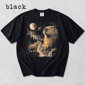 May include: Black t-shirt featuring a capybara illustration under a full moon in a forest setting. The image includes the word "black" at the top left. The design showcases a detailed depiction of the capybara, with a waterfall and trees.