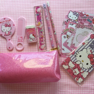 May include: Pink Hello Kitty themed pencil case with a zipper closure. The case is filled with Hello Kitty stationery including a ruler, pencils, a comb, an eraser, and stickers.