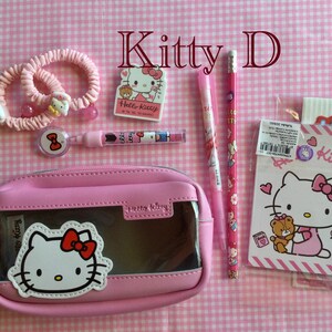 May include: Pink Hello Kitty themed stationery set with a pencil, pen, eraser, two hair ties, a pencil case, and two notebooks. The pencil case is clear and has a Hello Kitty face on the front. The notebooks have Hello Kitty faces on the front and say "Kitty" and "Hello Kitty" on the covers.