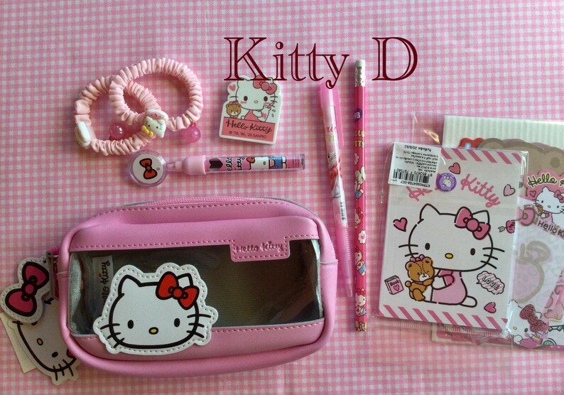 May include: Pink Hello Kitty themed stationery set with a pencil, pen, eraser, two hair ties, a pencil case, and two notebooks. The pencil case is clear and has a Hello Kitty face on the front. The notebooks have Hello Kitty faces on the front and say "Kitty" and "Hello Kitty" on the covers.